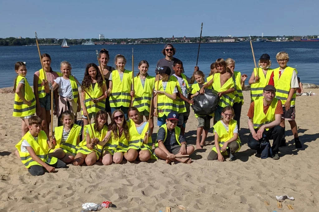 Gruppe von Kindern und Erwachsenen in Warnwesten mit Müllzangen bei einem CleanUp an einem Strand.