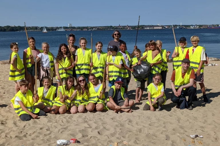 Gruppe von Kindern und Erwachsenen in Warnwesten mit Müllzangen bei einem CleanUp an einem Strand.