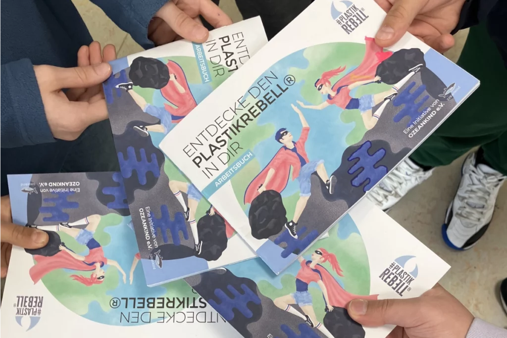 Kinder halten mehrere Exemplare des Arbeitshefts „Entdecke den Plastikrebell in dir“ in den Händen.