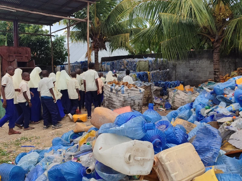 Schulkinder in Uniform besuchen auf Sansibar einen Recyclinghof mit großen Mengen gesammelter Plastikflaschen.