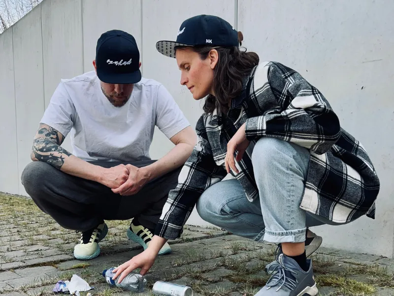 Zwei Erwachsene hocken an einer Wand und heben weggeworfenen Plastikmüll vom Boden auf.